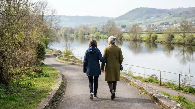 An older woman and her grown daughter walking arm in arm along a riverside path in early spring, seen from behind