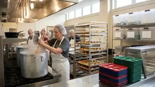 A busy stainless steel school kitchen with large pots on the stove, sheet pans of rolls and a long serving line, a cook in an apron and hairnet stirring a big pot